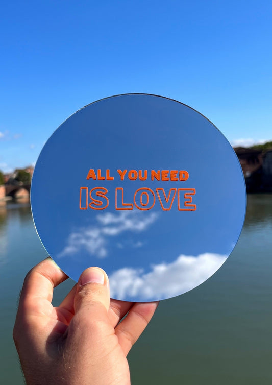 Miroir rond tenu dans une main avec la phrase All you need is love écrite en orange dessus. En fond se trouve le ciel bleu et la Garonne à Toulouse.