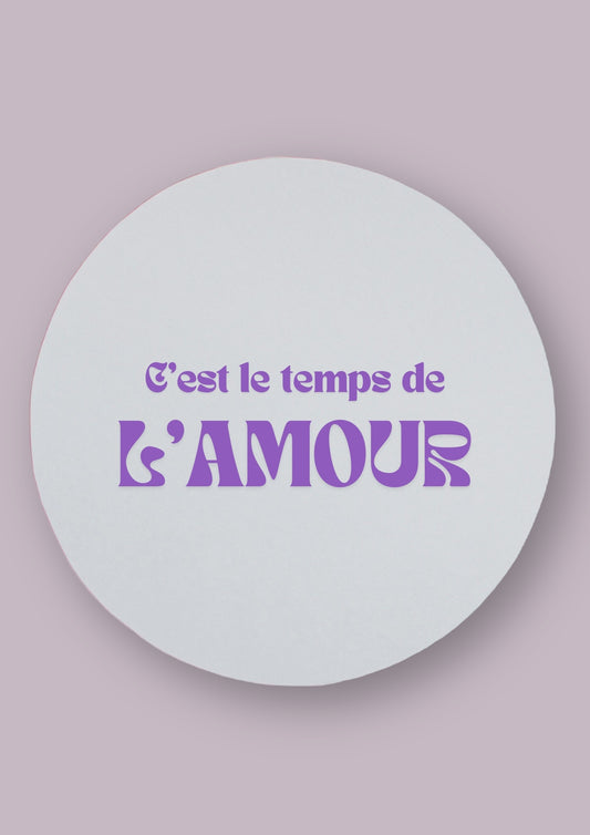Miroir rond avec la phrase c'est le temps de l'amour écrite en violet. Le fond de la photo est de couleur neutre. 