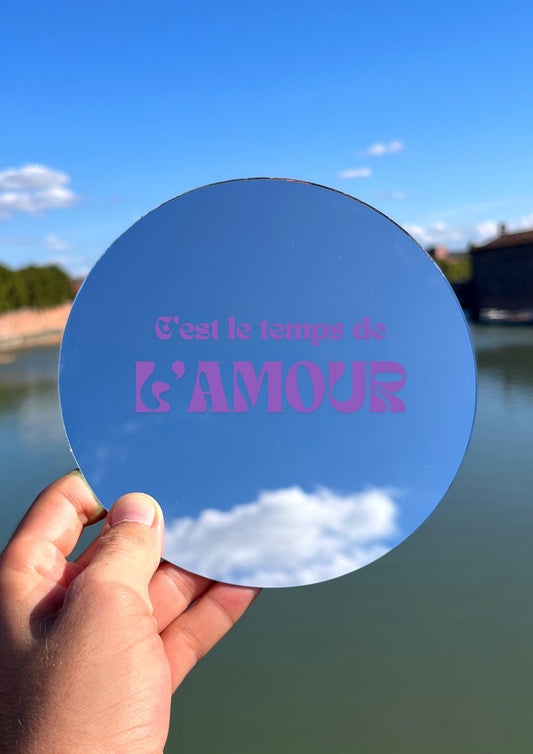 Miroir rond tenu dans une main avec la phrase c'est le temps de l'amour écrit en violet. En fond se trouve la Garonne de Toulouse. 
