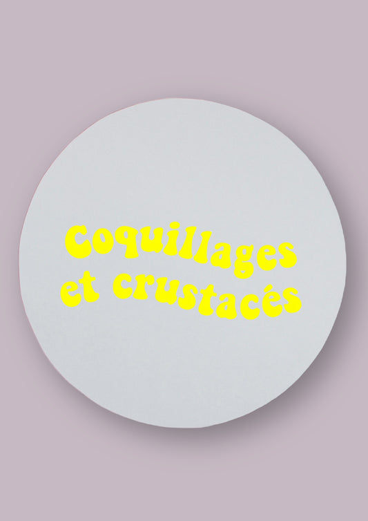 Miroir rond sur fond neutre, avec la phrase coquillages et crustacés écrit dessus en jaune.   