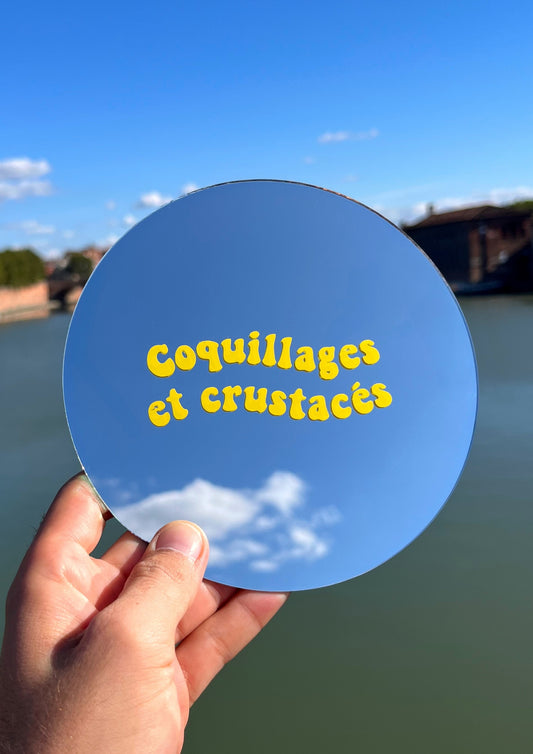 Miroir rond tenu dans une main avec la phrase coquillages et crustacés dessus. En fond se trouve la Garonne.  