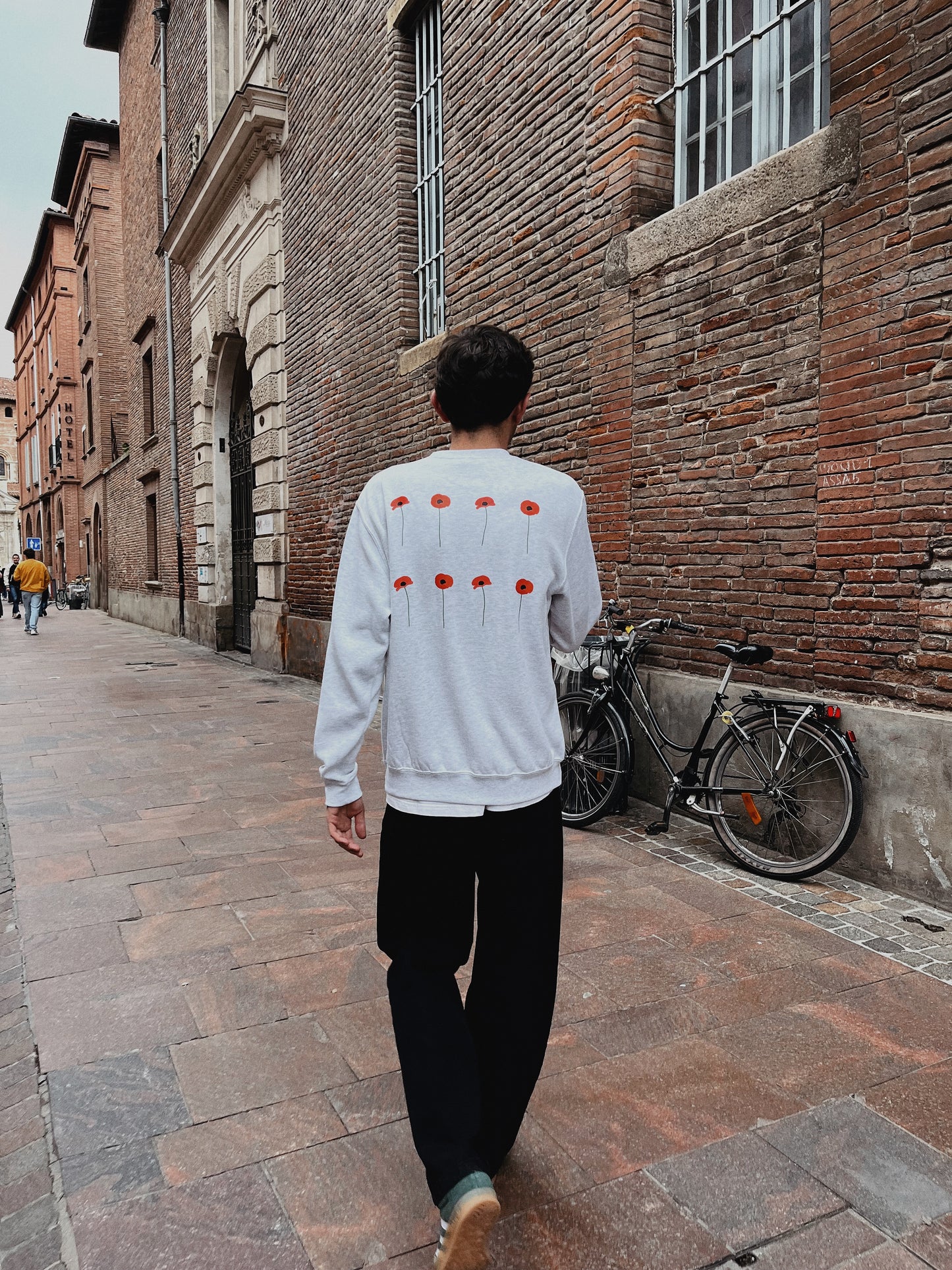 Jeune homme dos, pris en photo dans une rue toulousaine portant un sweat gris avec  coquelicots rouges sur le dos. 