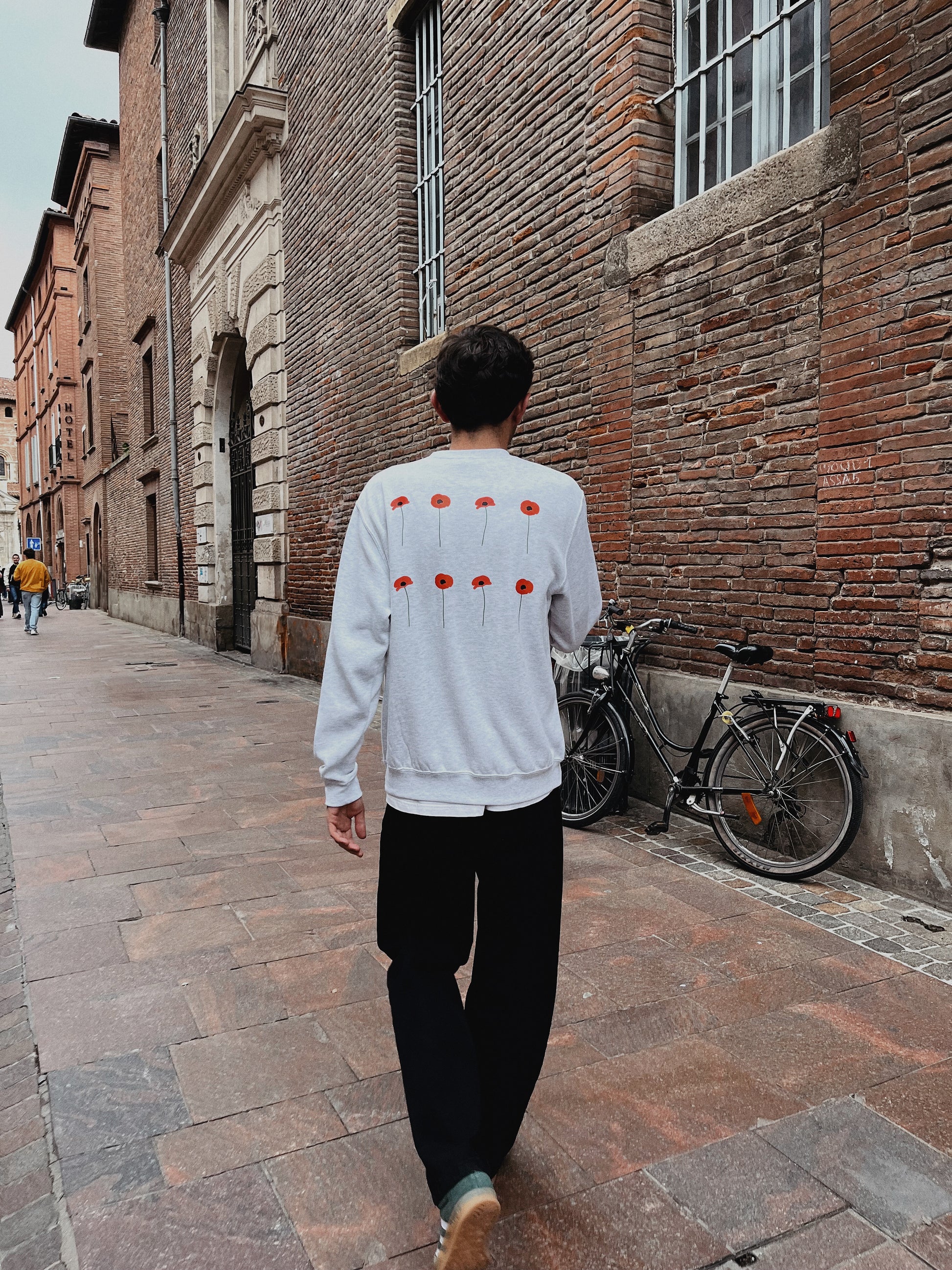 Jeune homme dos, pris en photo dans une rue toulousaine portant un sweat gris avec  coquelicots rouges sur le dos. 