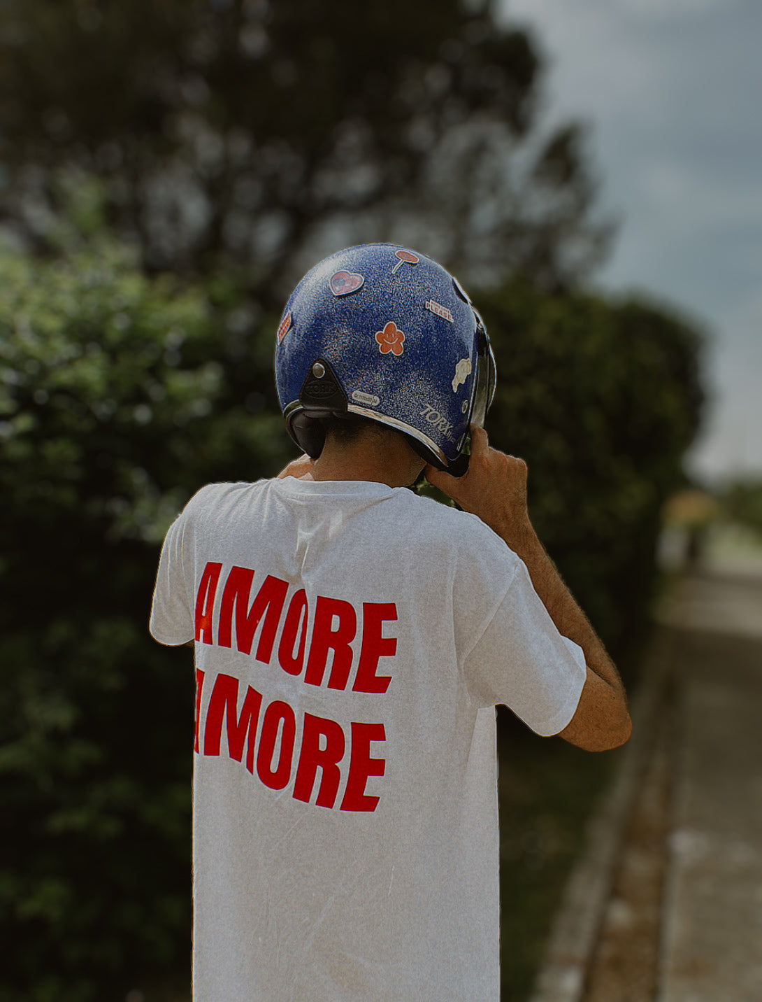 Photo d'un jeune homme de dos qui met un casque de moto bleu à paillettes sur la tête portant un t-shirt blanc avec l'inscription Amore Amore rouge dans le dos. 