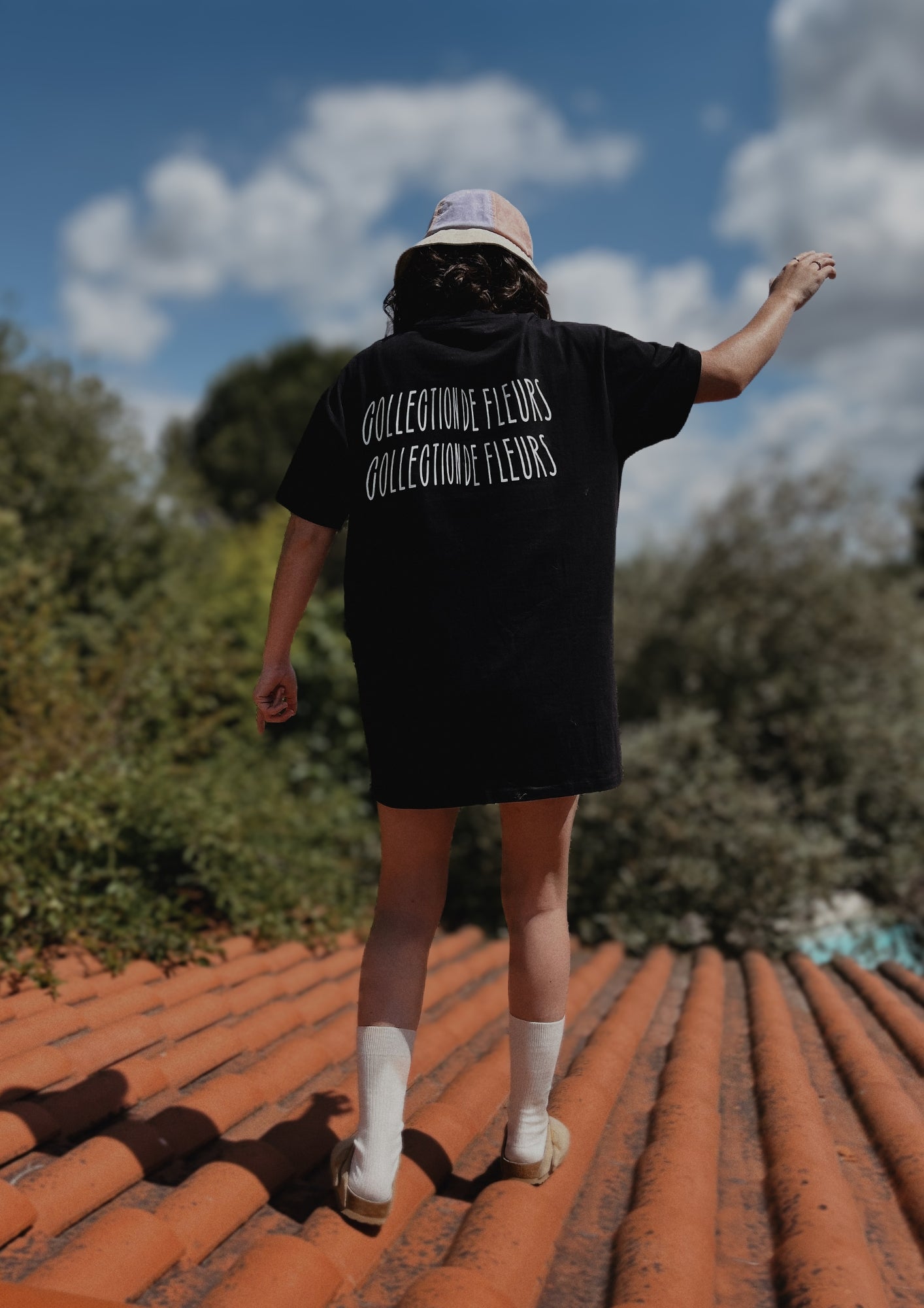 Jeune fille qui marche sur un toit de tuile orange en portant un t-shirt noir avec l'inscription "collection de fleurs"dans le dos. En fond des arbres et le ciel bleu.  