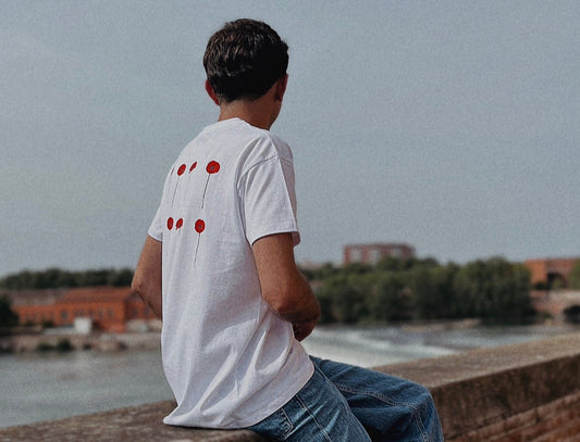 Photo d'un jeune homme de dos assis sur une murette regardant la Garonne. Il porte un t-shirt blanc avec des coquelicots dans le dos. 