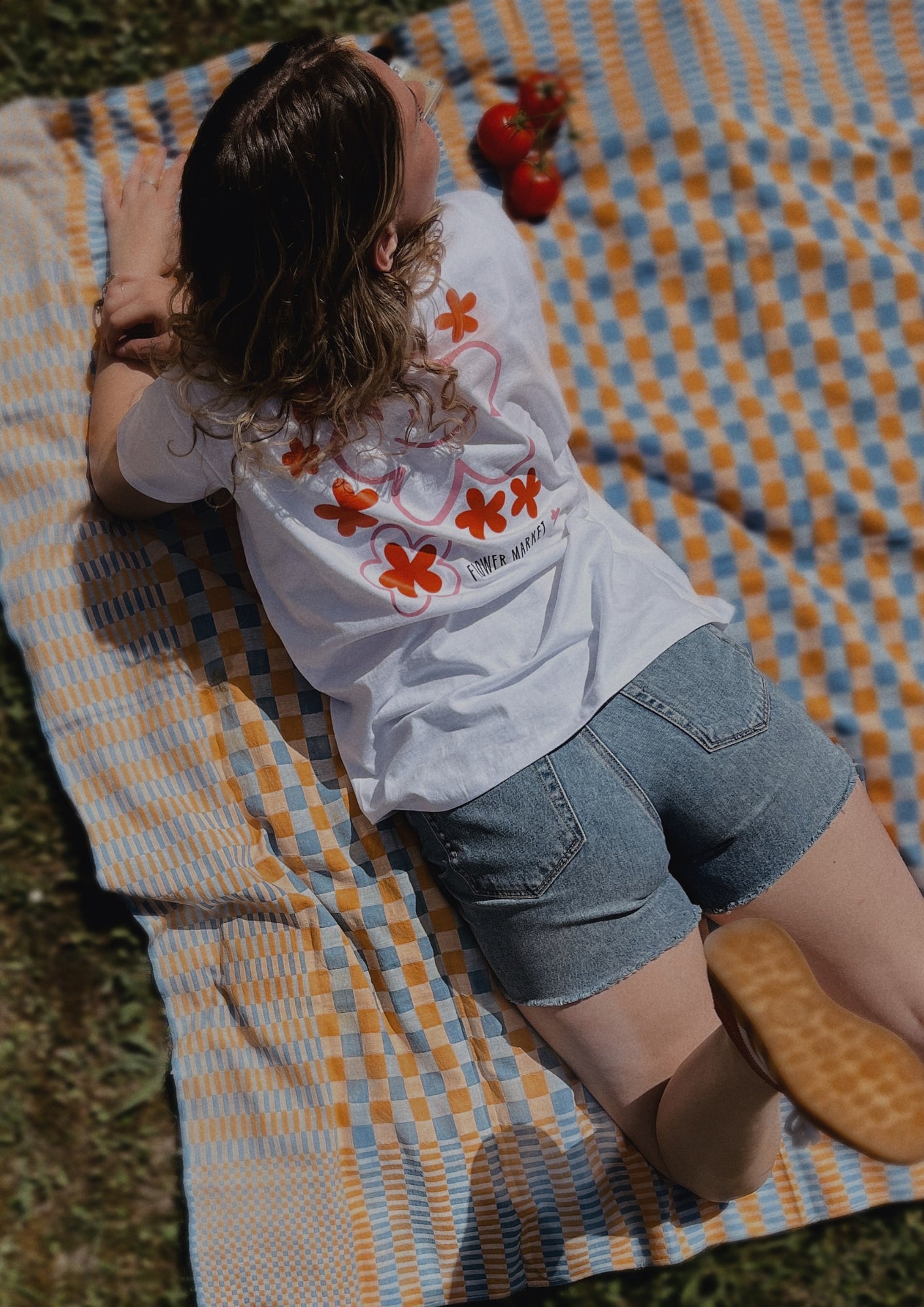 Jeune fille allongée sur le ventre sur une nappe à carreaux. Elle porte un t-shirt blanc avec une illustration de couleur rose et orange dans le dos. L'illustration représente des fleurs de différentes tailles, l'une d'entre elle à un smiley, et en bas à droite de l'illustration se trouve les mots flower market en noir. 