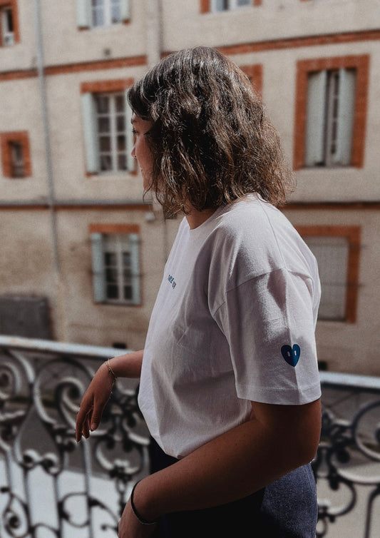 Mannequin femme sur un balcon qui pose de profil. Elle porte un t-shirt blanc avec l'inscription more amore sur le devant du t-shirt en bleue. Et un coeur bleue avec des yeux blancs sur la manche gauche. 