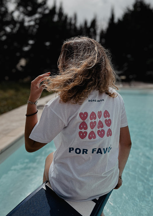 Photo d'une jeune fille de dos, assis sur un plongeoir avec une piscine et des arbres en fond. Elle porte un t-shirt blanc avec pour design la phrase More amor por favor en bleu et des coeurs roses. 